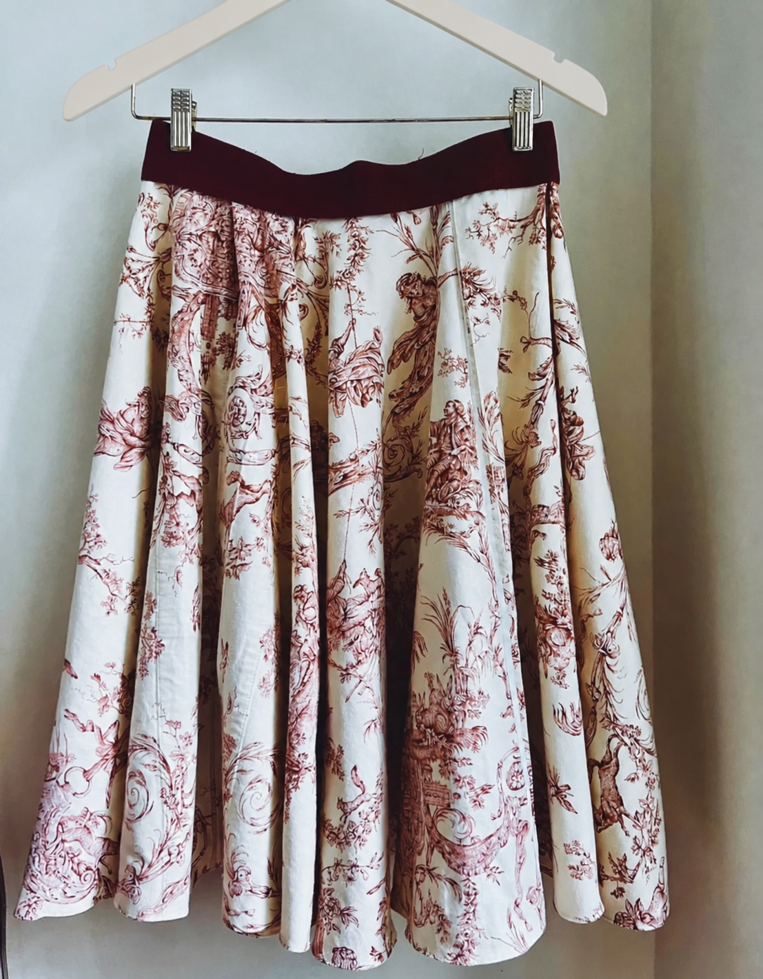 Finished cream and burgundy toile circle skirt hanging from a dark wooden hanger